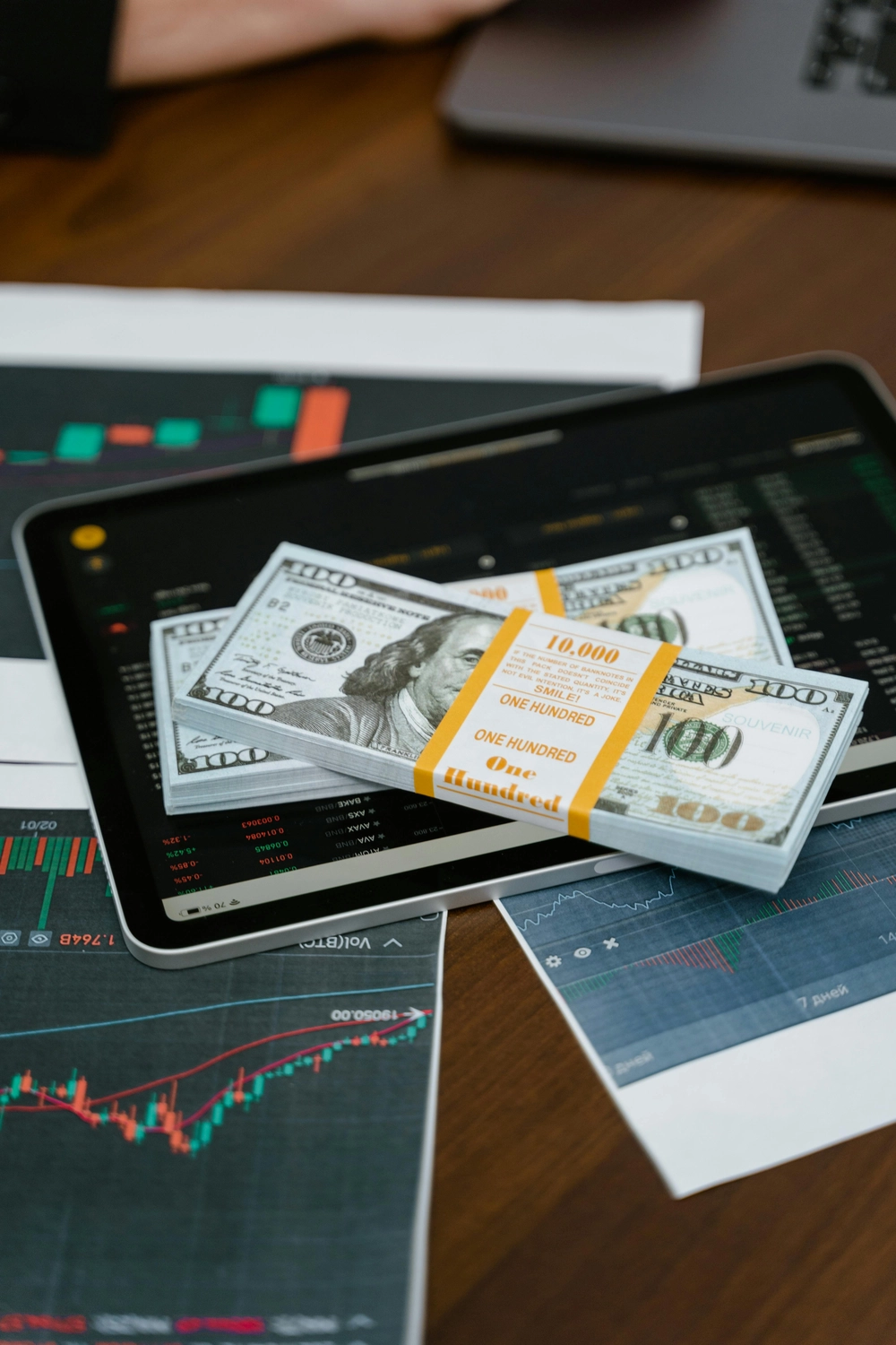 BIM - Bayerisches InvestManagement - InvestManagement - Stapel von US-Hundert-Dollar-Scheinen lagen auf einem Laptop, auf dem Finanzmarktdiagramme und -daten angezeigt wurden.