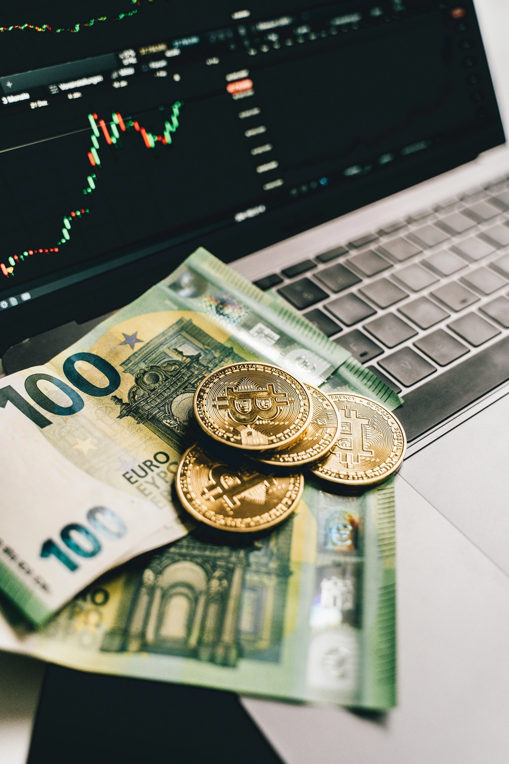 BIM - Bayerisches InvestManagement - InvestManagement - Eine 100-Euro-Banknote und goldfarbene Münzen, die wahrscheinlich Bitcoin repräsentieren, liegen auf einer Laptop-Tastatur, auf deren Bildschirm ein Finanzhandelsdiagramm angezeigt wird.
